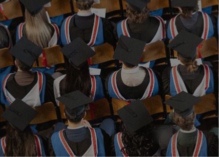 Overhead view of rows of graduates seated in an auditorium, wearing black hats and graduation gowns with blue, white, and red trim. Wooden seats are visible between the closely packed rows, creating a patterned scene of graduation caps and robes.